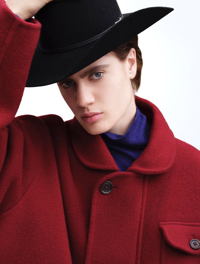 Portrait of a fair-skinned male model wearing a black hat and a red wool coat posing in a bright photo studio against a white background.
