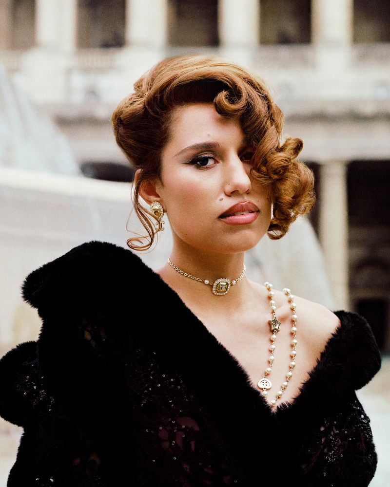Portrait of a woman with auburn curly hair, wearing gold jewellery and a black fur coat against a blurred architectural background.