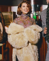 Smiling woman with wavy brown hair wearing a sheer embellished top and a fluffy cream coat in a bar setting.