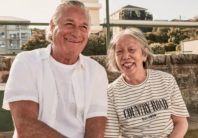 Elderly couple poses outdoors; the man wears a white button-down shirt, the woman wears a striped Country Road t-shirt, both smiling.