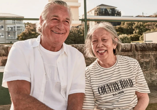 Elderly couple poses outdoors; the man wears a white button-down shirt, the woman wears a striped Country Road t-shirt, both smiling.