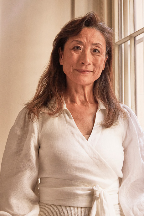 Close-up studio portrait of an adult woman with shoulder-length hair, wearing a white wrap top and smiling in front of a window.
