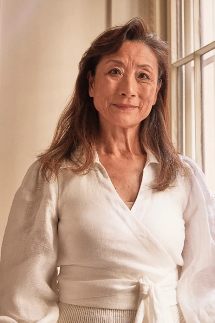 Close-up studio portrait of an adult woman with shoulder-length hair, wearing a white wrap top and smiling in front of a window.