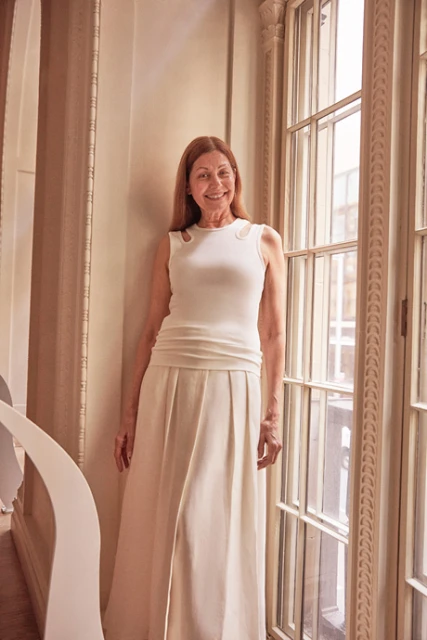 Elegant woman in a sleeveless white dress stands gracefully indoors near a window, illuminated by soft light. Fashion portrait.