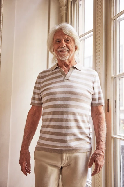 Portrait of an elderly man with long white hair standing by a window. He is wearing a striped shirt and beige pants, smiling gently.