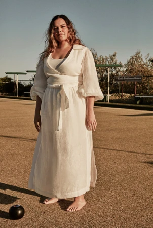 Woman in flowing white dress stands barefoot on a lawn bowling green in outdoor setting, bathed in sunlight, holding a bowling ball.