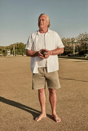 Mature man standing on a lawn bowling green holding a lawn bowling ball. He is wearing casual summer clothing on a sunny day.