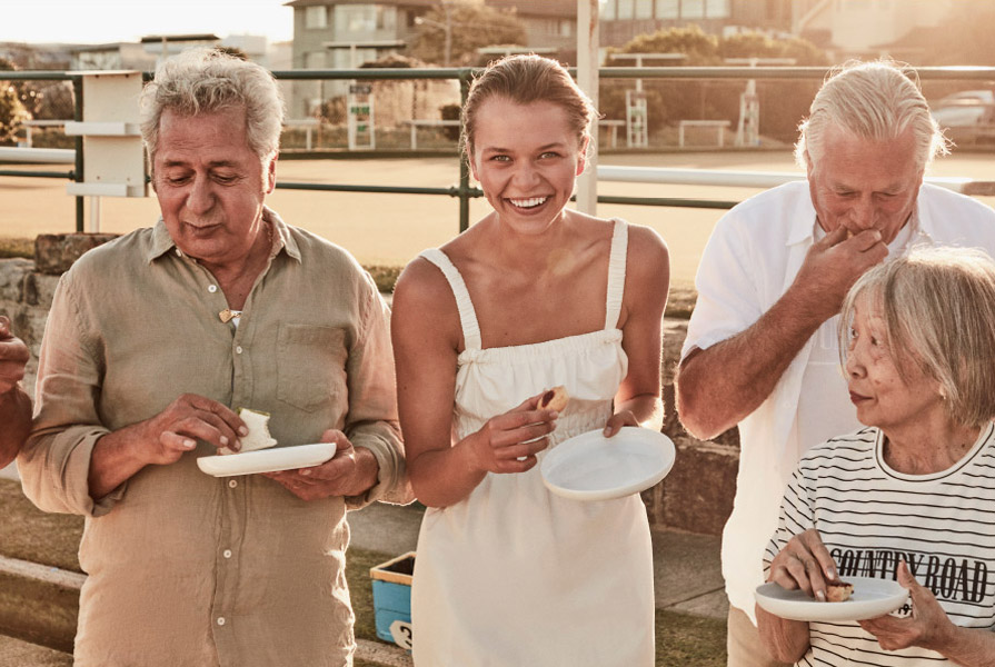 A group of adults are outside holding plates, smiling, and eating together in natural daylight. A casual social gathering.