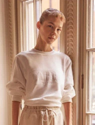 Model standing by a tall window wearing a white crewneck sweatshirt and light drawstring trousers.