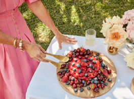 A woman in a pink dress arranges a wooden platter topped with a red berry dessert on a white tablecloth in a backyard setting.