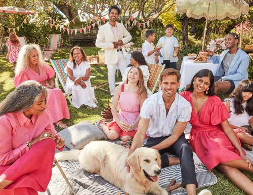 Outdoor garden party with a diverse group of people in pink and white outfits, enjoying the sunshine on the green grass.