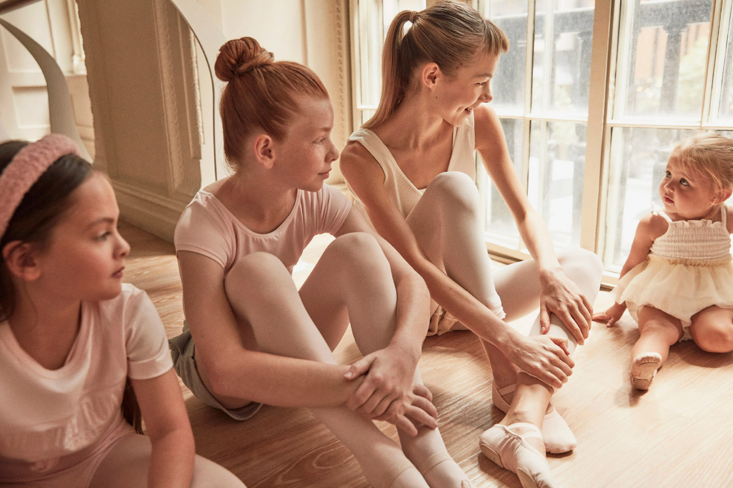 Four young ballerinas sit on a wood floor in a brightly lit room near a window, three in pink leotards and ballet slippers, one in a tutu.