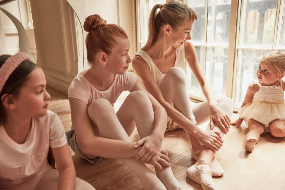 Four young ballerinas sit on a wood floor in a brightly lit room near a window, three in pink leotards and ballet slippers, one in a tutu.