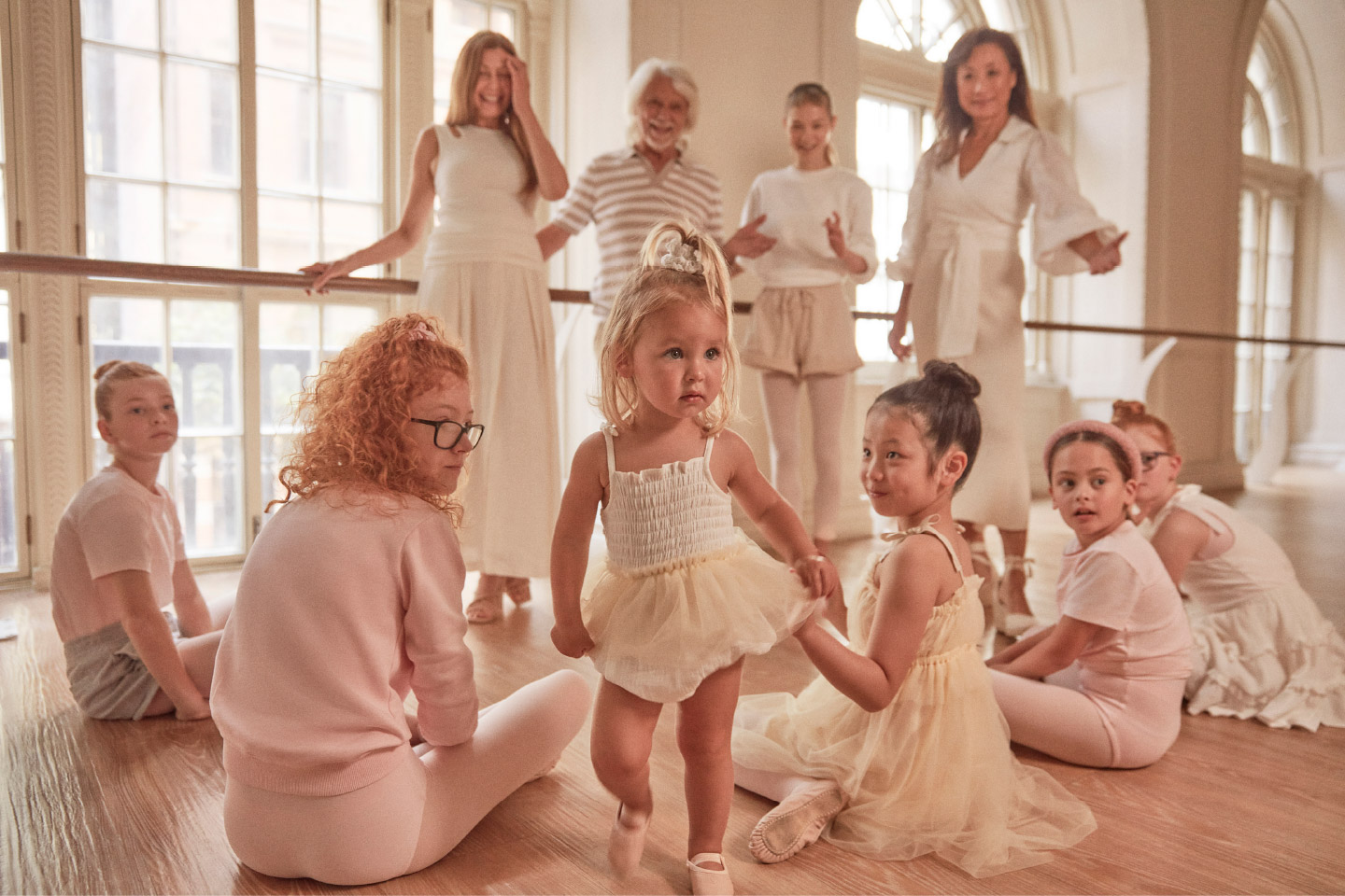 Group of children and adults in a bright dance studio, wearing light-colored clothes, gathered around a ballet barre on a wooden floor.