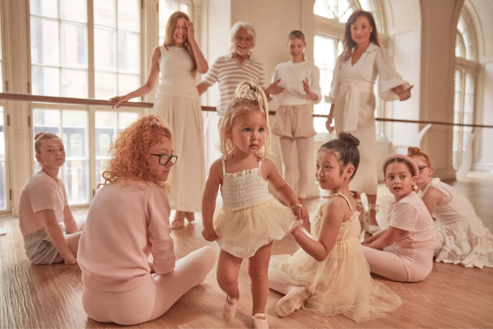 Group of children and adults in a bright dance studio, wearing light-colored clothes, gathered around a ballet barre on a wooden floor.