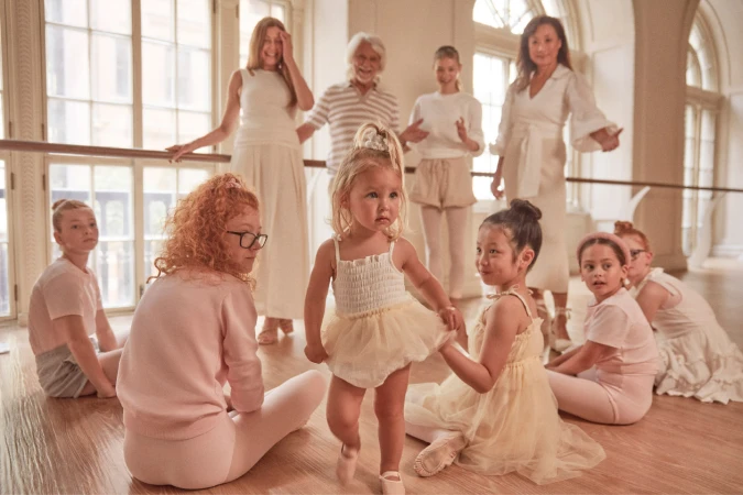 Group of children and adults in a bright dance studio, wearing light-colored clothes, gathered around a ballet barre on a wooden floor.
