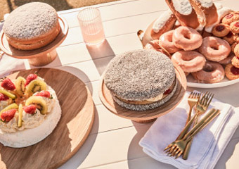 Overhead shot of assorted cakes and pastries on a white table, including fruit cake and sugar-dusted pastries, with gold forks. 