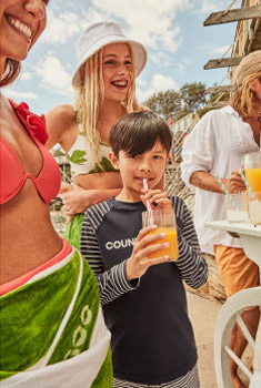 A young boy in a striped shirt drinks orange juice with a straw. Beachside scene with adults and white table in the background.