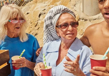 Three people, two elderly women with beach towel on head, one young man, holding drinks, on a sunny beach with cliffs backdrop.