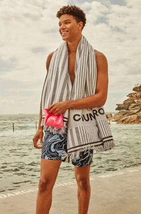 Young man smiles on a sunny beach, wearing swim trunks and a striped towel, with the ocean and rocky cliffs in the background.