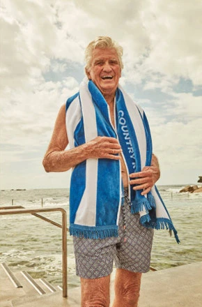 Elderly man smiles at beach, wearing patterned swim trunks and a blue-and-white towel draped around neck, with ocean background.