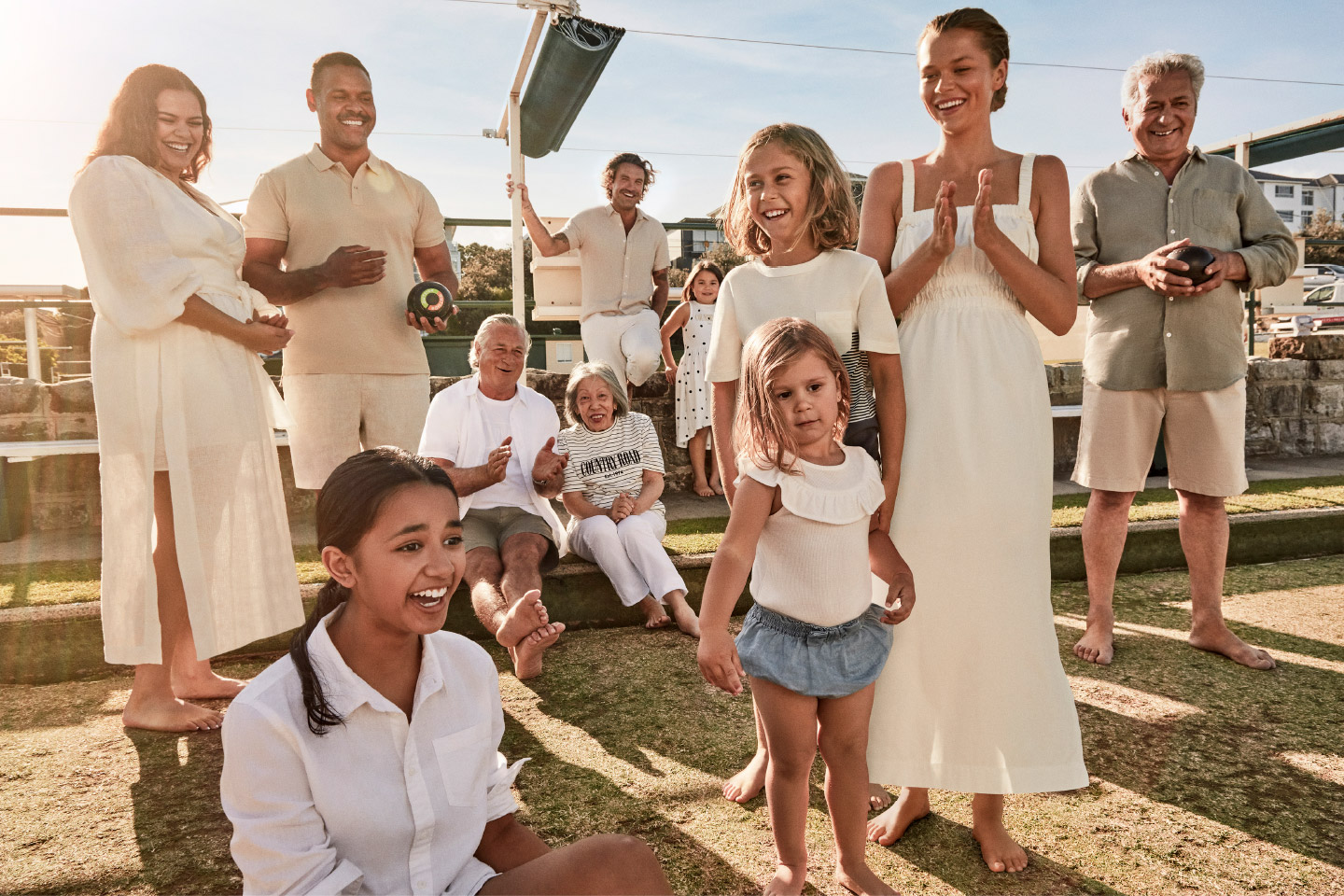 Diverse group of people, including adults and children, gathered on a grassy lawn with lawn bowls and residential buildings in the background.