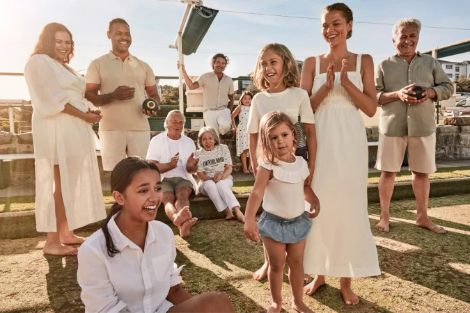 Diverse group of people, including adults and children, gathered on a grassy lawn with lawn bowls and residential buildings in the background.