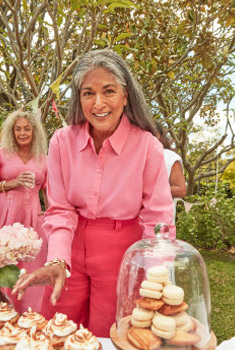 An elderly woman in a pink outfit smiles at an outdoor garden party with a cake stand, a gathering of people, and bright, natural light.