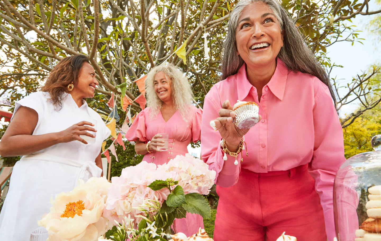 Three mature women enjoy an outdoor party with cupcakes, flowers, and decorative flags, dressed in pastel and white outfits, smiling.