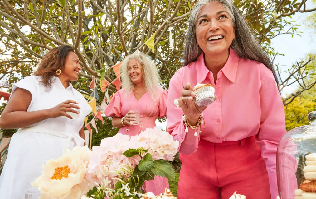 Three mature women enjoy an outdoor party with cupcakes, flowers, and decorative flags, dressed in pastel and white outfits, smiling.