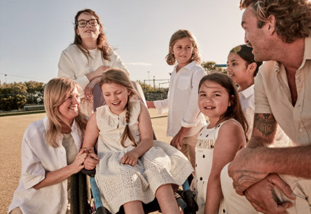 Smiling family group of 7 outdoors with a young girl with a disability using a wheelchair, posing in daytime light. 