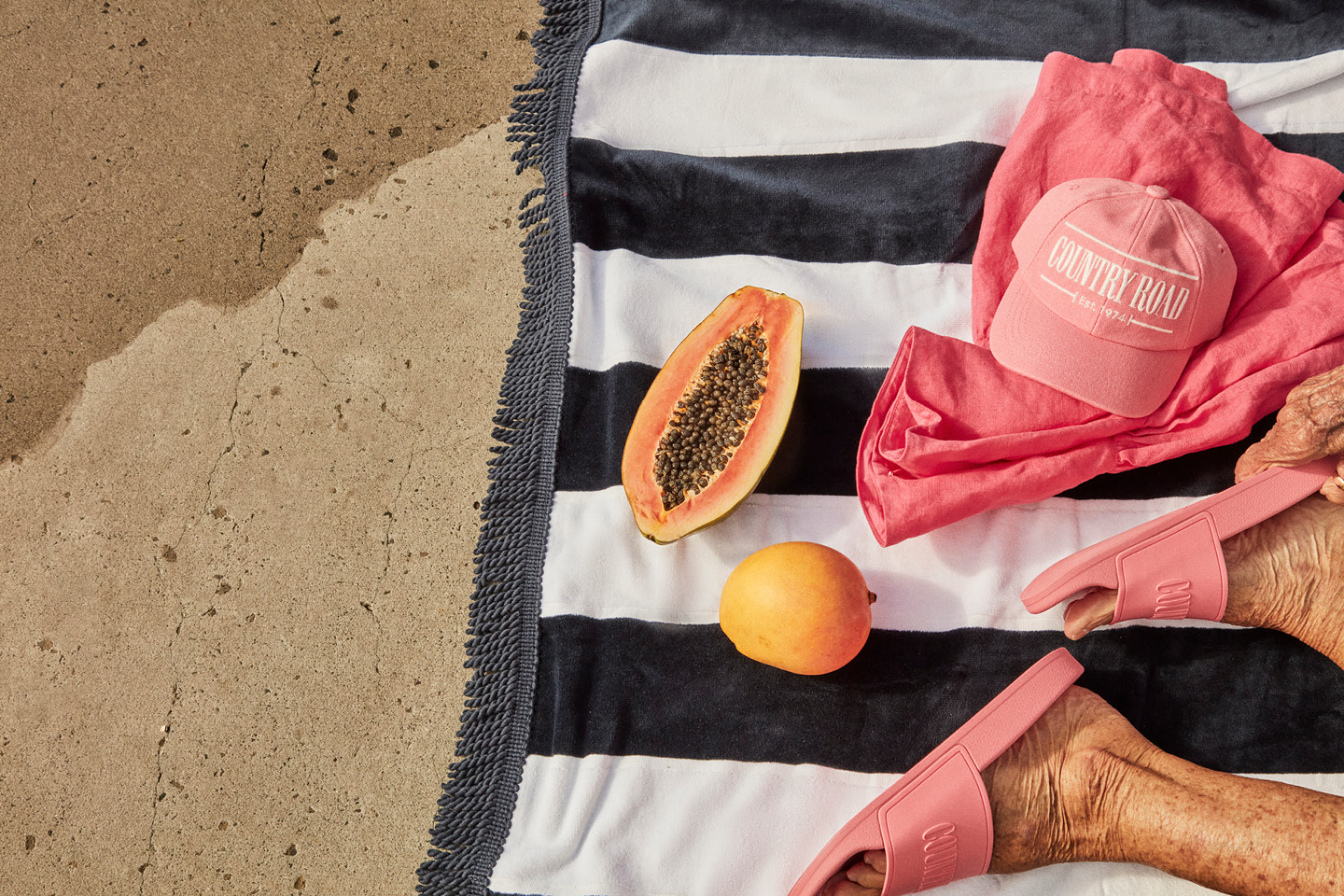 Overhead shot of a beach towel with a halved papaya, mango, baseball cap, apparel, and slide sandals, on sand, bathed in sunlight.