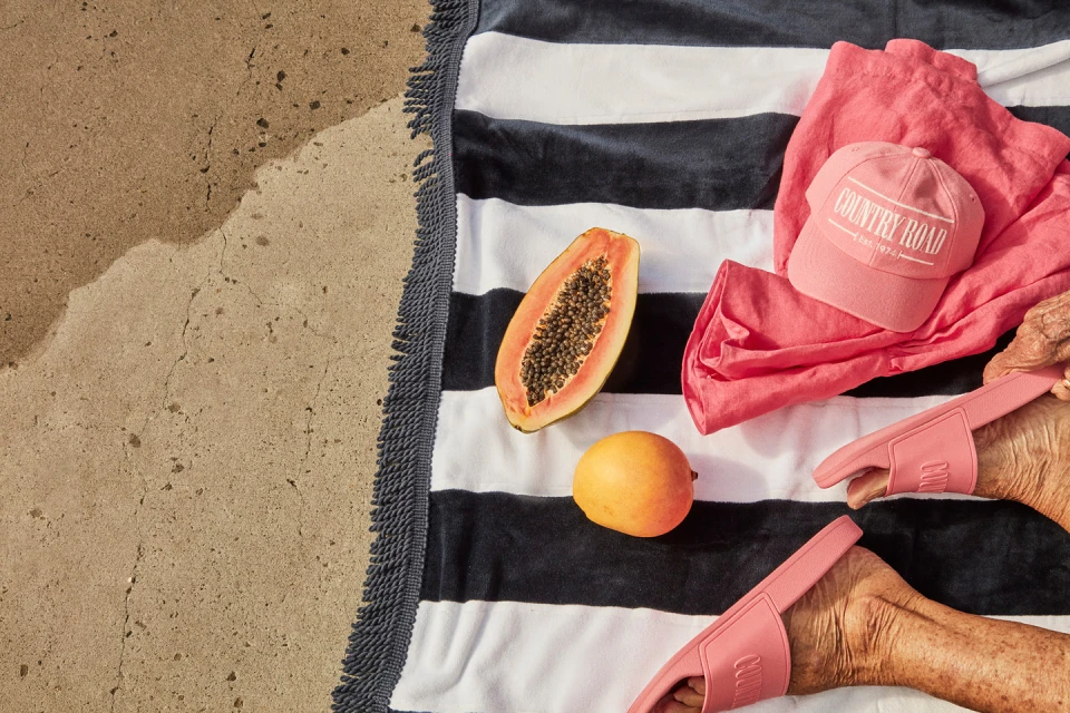 Overhead shot of a beach towel with a halved papaya, mango, baseball cap, apparel, and slide sandals, on sand, bathed in sunlight.