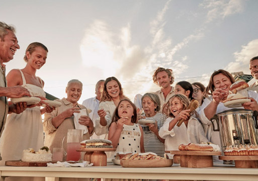 A diverse group of people, including children and adults, gather outdoors around a table filled with various dishes, creating a sense of community.