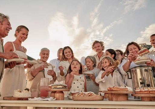 A diverse group of people, including children and adults, gather outdoors around a table filled with various dishes, creating a sense of community.