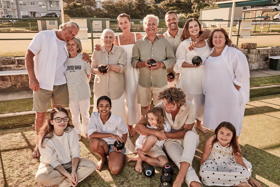 Large multigenerational group of fourteen people posing on a lawn bowling green, wearing light-colored clothing, holding lawn bowling balls.