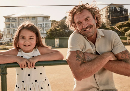Father and young daughter posing outdoors on a sunny day, smiling and leaning against a green rail. Man with arm tattoos.