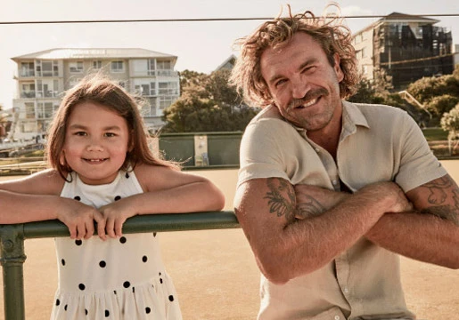 Father and young daughter posing outdoors on a sunny day, smiling and leaning against a green rail. Man with arm tattoos.