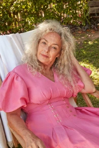Elegant older woman with silver hair and a pink dress sitting in a wooden chair outside on a sunny day in a natural setting.