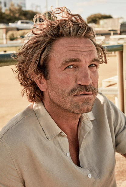 Portrait of an adult male with wavy auburn hair, a mustache, and a light tan shirt against a neutral outdoor background.