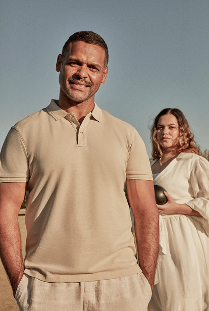 Portrait of a man in a beige polo shirt with a woman in a white dress holding a ball; they are standing outdoors on a sunny day.