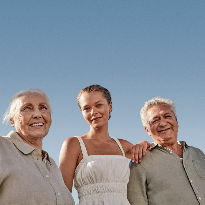 Three models including an older woman, a younger woman in a white dress, and an older man against a clear blue sky