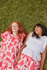 Two young girls lying on green grass outdoors, smiling and wearing colorful patterned dresses.