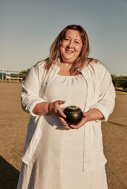 Smiling woman holding a lawn bowl on a lawn bowling green, wearing a white dress and shirt, outdoors on a sunny day with blue sky.