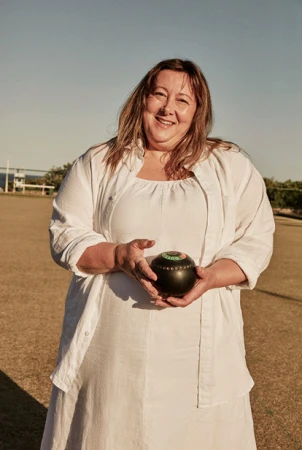 Smiling woman holding a lawn bowl on a lawn bowling green, wearing a white dress and shirt, outdoors on a sunny day with blue sky.