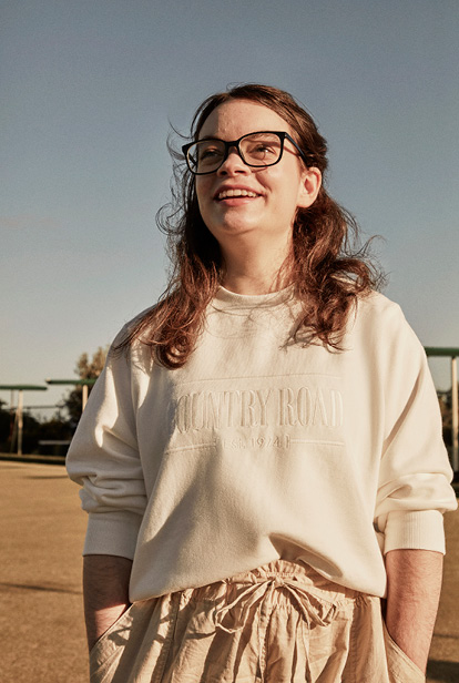 Smiling young adult female wearing glasses, a cream-colored sweatshirt, and beige drawstring pants, standing outdoors on a sunny day.