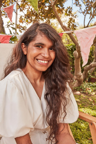 A smiling woman with long, dark curly hair sits outside in a white dress with puffy sleeves in front of trees and party banners.