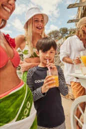 A young boy drinking juice through a pink straw surrounded by smiling family members outdoors for Country Road.