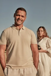 Man in beige polo shirt standing outdoors with a woman holding an object in the background for Country Road