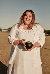 Smiling woman in white shirt holding a black lawn bowl on a bowling green outdoors